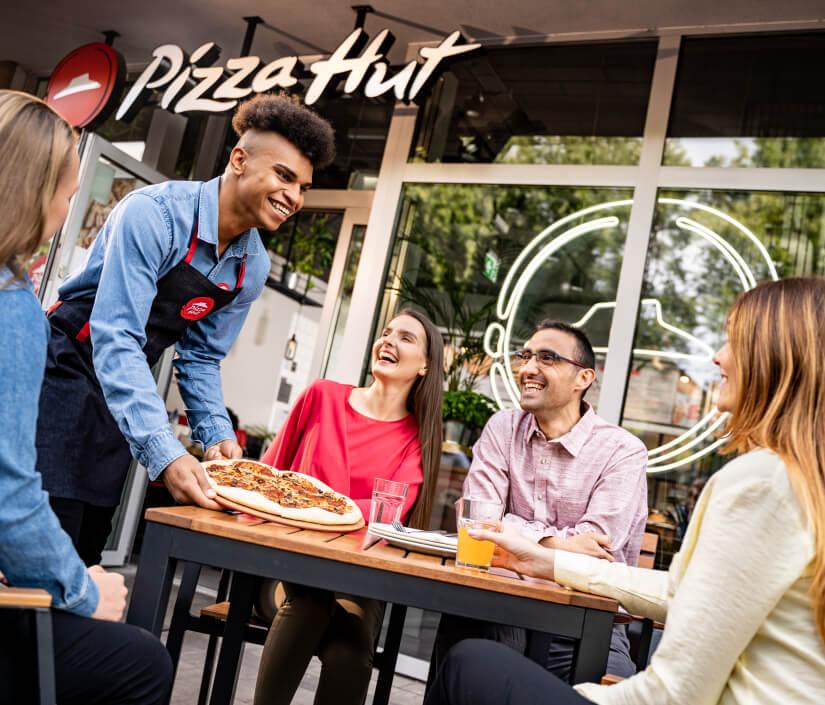 Happy group of friends enjoying fresh pizza served by smiling waiter at an outdoor Pizza Hut restaurant